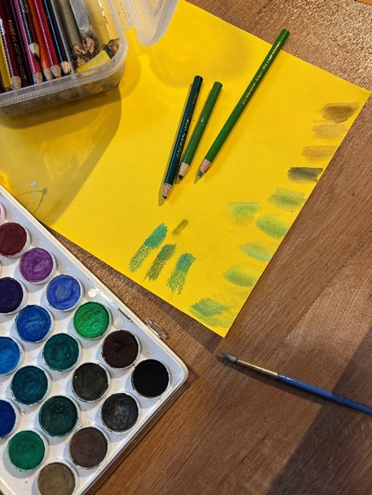 Green colored pencils on top of yellow paper next to a bin of colored pencils and watercolor paints.